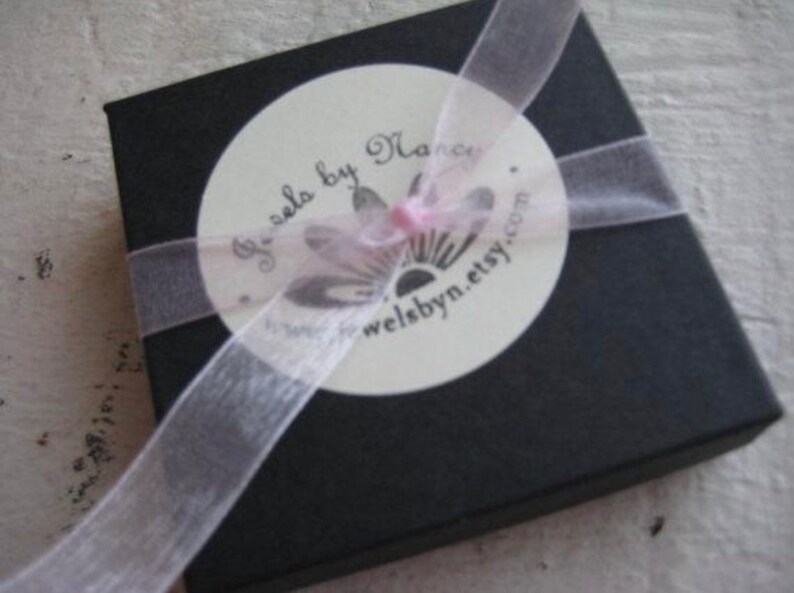 May include: A black square gift box with a white circular label and a sheer ribbon. The label reads "Jewels by Nancy" and "welsbyn.etsy.com". The ribbon is wrapped around the box and tied in a bow.
