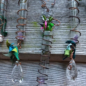 Pode incluir: Três tubos de vidro envoltos em fio de cobre, cada um com um cristal na parte inferior e um elemento decorativo na parte superior. Os tubos estão pendurados em uma superfície de madeira. Os elementos decorativos incluem uma flor, uma flor de tecido azul e verde e uma flor de tecido verde e branco.