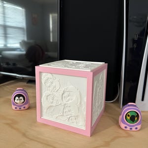 May include: A square lamp with a pink frame and white panels featuring embossed portraits of multiple faces on each side. Two small purple and pink portable speakers, one with a penguin and one with a parrot, sit on either side of the lamp.