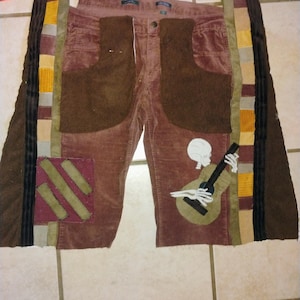 Puede incluir: Pantalón de pana marrón con detalles de patchwork. El pantalón tiene un esqueleto tocando una guitarra en una pierna y un parche burdeos con tres rayas en la otra pierna.