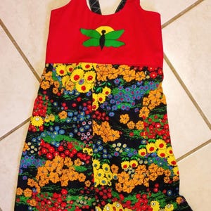 May include: A red sundress with a butterfly design and a floral skirt. The top features a butterfly design with green wings and a yellow sun. The skirt has a black background with colorful flowers.