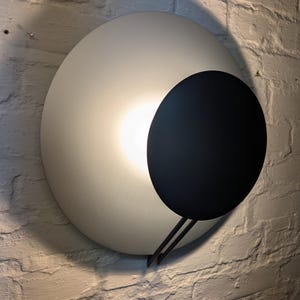 Peut inclure: Une applique murale moderne avec un abat-jour circulaire blanc et un accent circulaire noir. La lumière est allumée et éclaire la pièce.