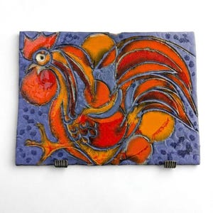 May include: A rectangular ceramic wall tile featuring a stylized rooster in vibrant shades of orange and red against a textured periwinkle blue background. The rooster design includes bold dark outlines and circular orange accents.
