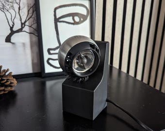 May include: A black speaker with a silver and black speaker head. The speaker is on a black table with framed art in the background. A black power cord extends from the speaker.