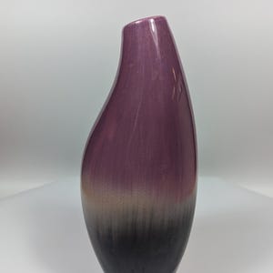 Könnte beinhalten: Eine hohe, tropfenförmige Vase mit Farbverlauf. Die Oberseite ist tiefviolett, geht in einen graubraunen Mittelteil über und endet in einem schwarzen Sockel. Die Vase hat eine glatte, glänzende Oberfläche und eine leicht abgewinkelte Öffnung.