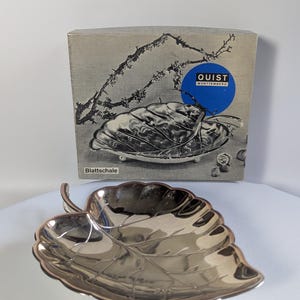 May include: A silver-colored leaf-shaped dish with detailed veining, likely for serving or display. The dish is in front of a box with the text "QUIST WURTTEMBERG" and the German word "Blattschale".