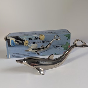 Puede incluir: Un abridor de botellas cromado con forma de delfín, con cola curvada y aleta. El abridor está junto a una caja con las palabras "Delphin Flaschenöffner" y una ilustración de una botella.