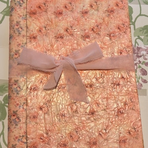 Pode incluir: Um diário decorativo com um padrão floral em tons de pêssego e rosa. A capa tem uma aparência texturizada e é amarrada com uma fita rosa claro. As bordas do diário também são decoradas com um padrão floral.