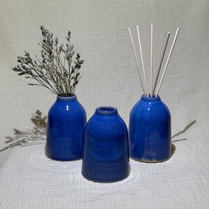 May include: Three cobalt blue ceramic vases. One vase holds dried flowers, another has wooden sticks, and the third is empty. The vases have a glossy finish and a rounded shape, ideal for home decor.