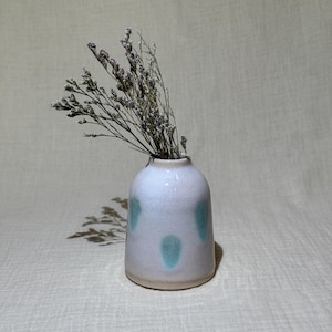 May include: A small, white ceramic vase with three turquoise spots. The vase contains a bundle of dried, purple-toned flowers. The vase has a rounded shape and a slightly textured surface, creating a rustic aesthetic.