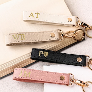May include: Four personalised keyrings in cream, tan, black, and pink. Each keyring features gold-coloured hardware and is embossed with initials. The keyrings are arranged on a neutral background with a book and paper.