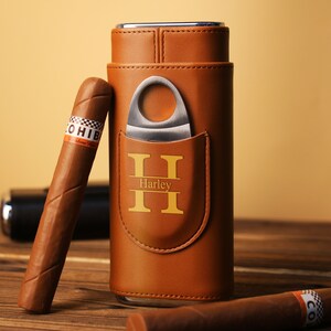 K&ouml;nnte beinhalten: Ein braunes Leder-Zigarrenetui mit integriertem Cutter und einem goldenen Monogramm "H" mit dem Namen "Harley" darunter. Zwei Zigarren sind neben dem Etui abgebildet.
