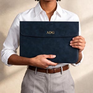Peut inclure: Une housse pour ordinateur portable rectangulaire bleu marine avec une finition lisse et mate et une fermeture à rabat. Le centre du rabat présente les lettres ADG dorées en relief. La housse a des coins arrondis et un design structuré.
