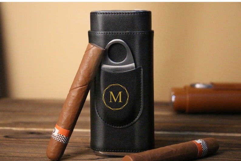 K&ouml;nnte beinhalten: Schwarze Leder-Zigarrenh&uuml;lle mit integriertem Cutter und goldenem Monogramm "M". Zwei braune Zigarren liegen neben der H&uuml;lle.