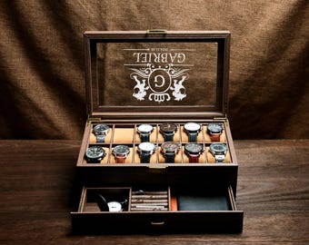 May include: A wooden watch box with a glass top and a drawer. The box is personalised with the inscription "GABRIEL" and a crest featuring the letter "G". The box has 12 slots for watches and a drawer for accessories.