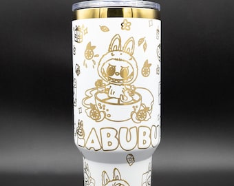 May include: White tumbler with gold accents and a clear straw. The tumbler features a repeating pattern of cartoon bunnies and the word "ABUBU" in gold. The lid and rim are gold.