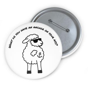 May include: A round white pin-back button featuring a black line drawing of a sheep wearing sunglasses with an anchor symbol on its side. The text "WHAT DO YOU HAVE AN ANCHOR ON YOUR ASS?" is printed in black along the top edge of the button.