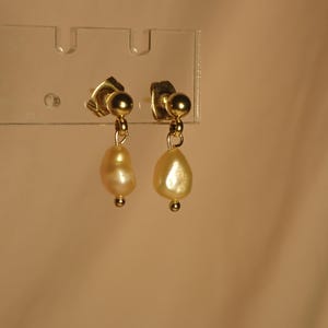 May include: A pair of gold earrings featuring light-yellow, pear-shaped pearls. The earrings are presented on a clear plastic holder. Each earring has a small gold ball at the top and bottom of the pearl.