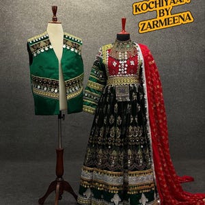 Puede incluir: Un vestido negro y verde con bordados dorados y un chal rojo. El vestido tiene un cuello alto y mangas largas. El chal está drapeado sobre los hombros y tiene un ribete de flecos. El vestido está en un maniquí. El texto "KOCHIYYAAN BY ZARMEENA" está en la esquina superior derecha de la imagen.