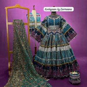 Może przedstawiać: Turkusowo-niebieska haftowana sukienka ze srebrnymi detalami, prezentowana na manekinie. W pobliżu jest drapowana pasująca szal. Obraz zawiera tekst "Kochiyaan by Zarmeena".