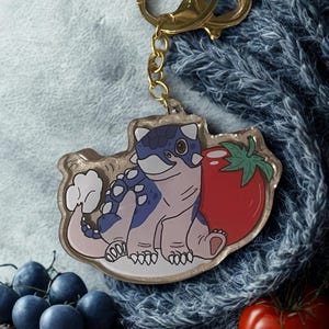 Puede incluir: Un llavero dorado con un dinosaurio de dibujos animados con manchas azules y blancas, sentado junto a un tomate rojo. El llavero está sujeto a un cierre y una cadena dorados.