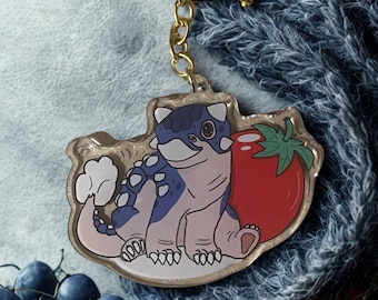 May include: A gold-coloured keyring featuring a cartoon dinosaur with blue and white spots, next to a red tomato. The keyring is attached to a gold-coloured clasp and chain.