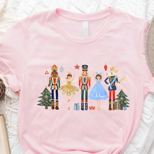 May include: Pink t-shirt with a colorful illustration of the Nutcracker and other characters from the ballet. The characters are standing in front of a Christmas tree.