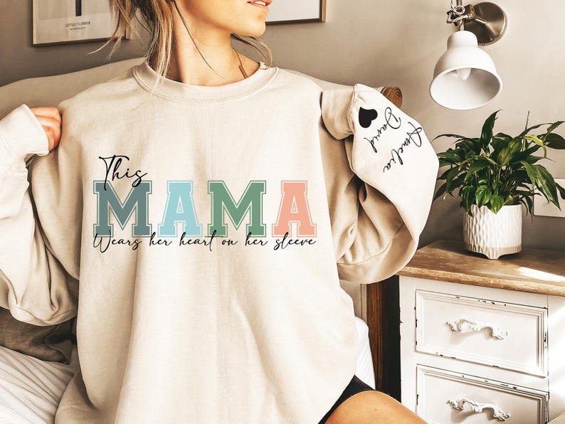 May include: A beige sweatshirt with the text "This Mama wears her heart on her sleeve" in colorful letters. The sleeve has a black heart with the text "Forever Loved" in black.