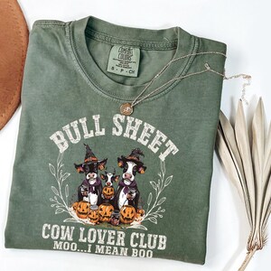 May include: An olive green crew neck t-shirt featuring a graphic of three cows wearing witch hats surrounded by pumpkins. The text reads BULL SHEET COW LOVER CLUB MOO...I MEAN BOO. The shirt has a Comfort Colors label indicating size small.
