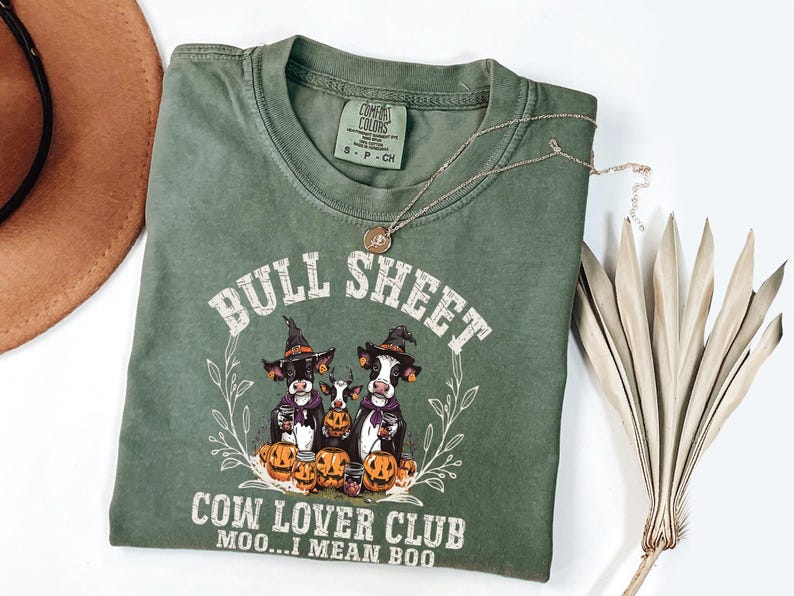 May include: An olive green crew neck t-shirt featuring a graphic of three cows wearing witch hats surrounded by pumpkins. The text reads BULL SHEET COW LOVER CLUB MOO...I MEAN BOO. The shirt has a Comfort Colors label indicating size small.