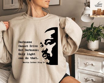 Może przedstawiać: Beżowa bluza z cytatem "Darkness Cannot Drive Out Darkness; Only light can do that." i czarną sylwetką Martina Luthera Kinga Jr. Tekst i obraz znajdują się z przodu ubrania.