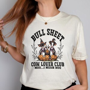 May include: A cream-colored t-shirt featuring a graphic of three cows wearing witch hats and holding pumpkins. The text reads BULL SHEET COW LOVER CLUB MOO...I MEAN BOO. The design is framed by black line-art leaves and vines on a light background.