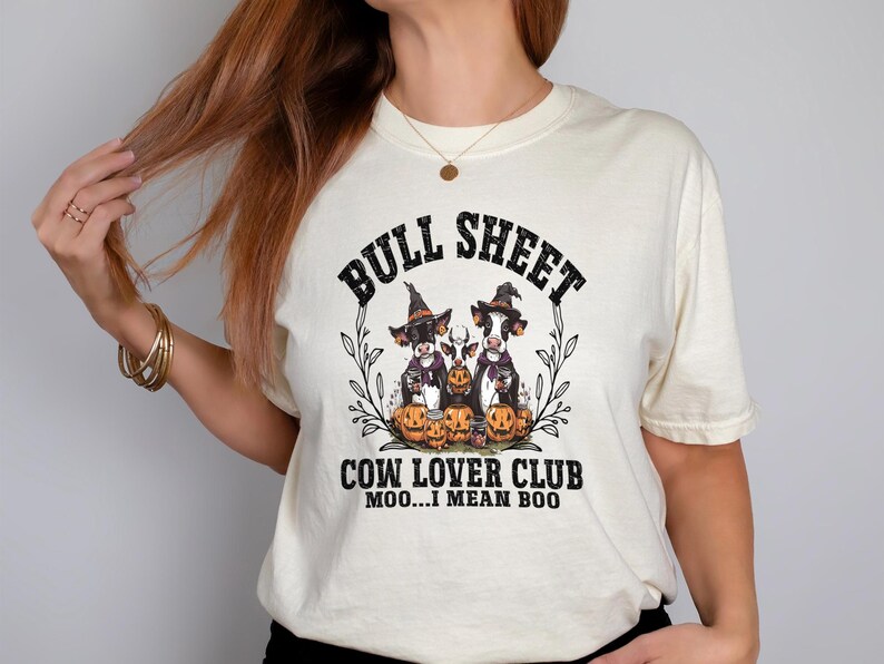 May include: A cream-colored t-shirt featuring a graphic of three cows wearing witch hats and holding pumpkins. The text reads BULL SHEET COW LOVER CLUB MOO...I MEAN BOO. The design is framed by black line-art leaves and vines on a light background.