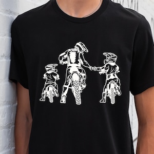 May include: A black t-shirt with a white graphic of three people riding dirt bikes. The people are wearing helmets and goggles. The graphic is in a silhouette style.