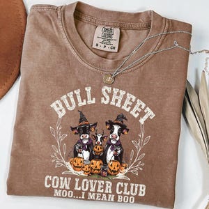 May include: A brown short-sleeve t-shirt featuring a graphic of three cows wearing witch hats surrounded by pumpkins. The text reads BULL SHEET COW LOVER CLUB MOO...I MEAN BOO. The shirt has a Comfort Colors label at the neck and a crew neckline.