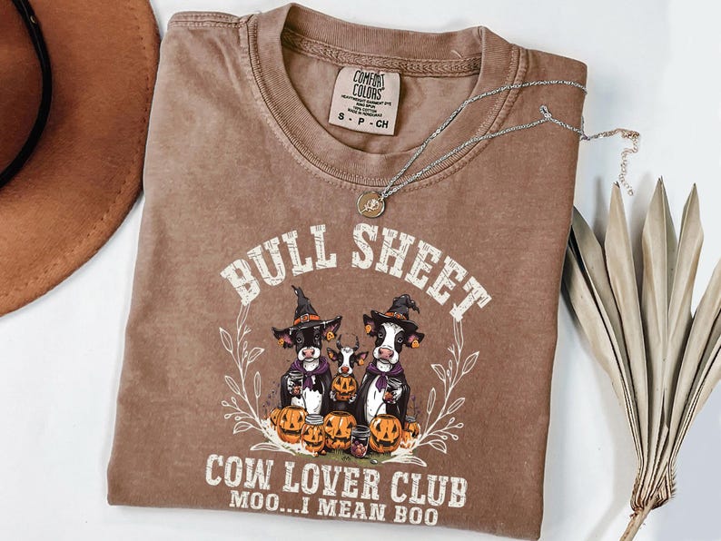 May include: A brown short-sleeve t-shirt featuring a graphic of three cows wearing witch hats surrounded by pumpkins. The text reads BULL SHEET COW LOVER CLUB MOO...I MEAN BOO. The shirt has a Comfort Colors label at the neck and a crew neckline.
