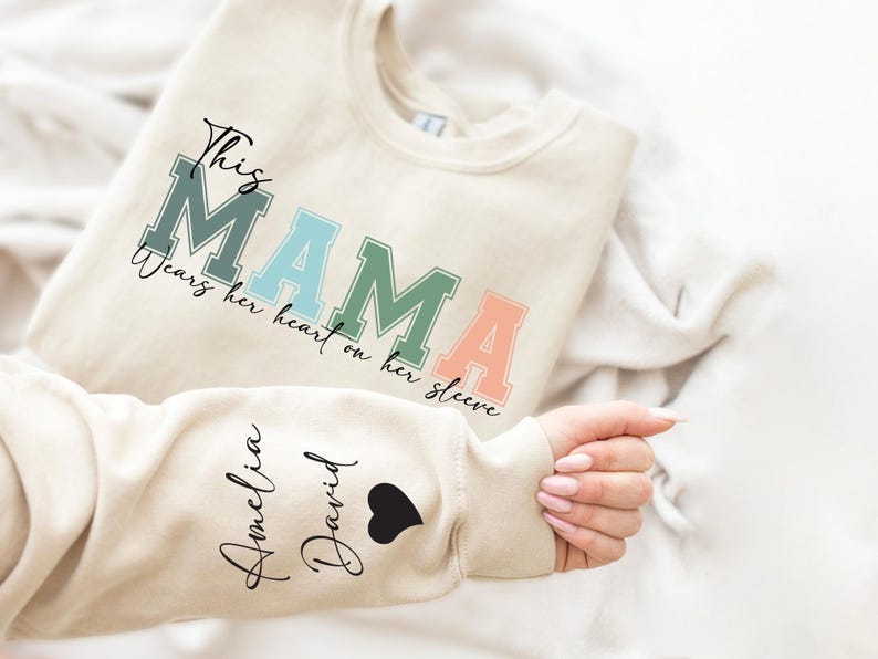 May include: A beige sweatshirt with the text "This Mama wears her heart on her sleeve" in rainbow colors. The sleeve has the names "Amelia" and "David" written in black cursive with a black heart.
