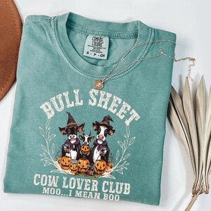May include: A sage green short-sleeve t-shirt featuring a graphic of three cows wearing witch hats surrounded by pumpkins. The text reads BULL SHEET COW LOVER CLUB MOO...I MEAN BOO. The shirt has a Comfort Colors label at the neck and a crew neckline.
