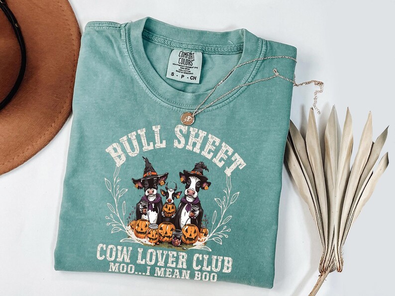 May include: A sage green short-sleeve t-shirt featuring a graphic of three cows wearing witch hats surrounded by pumpkins. The text reads BULL SHEET COW LOVER CLUB MOO...I MEAN BOO. The shirt has a Comfort Colors label at the neck and a crew neckline.