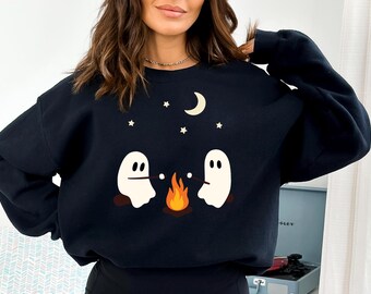 May include: A black crewneck sweatshirt featuring a graphic print of two white ghosts sitting by a campfire roasting marshmallows. Above the ghosts, there is a crescent moon and several small stars in a light cream color against the dark fabric.