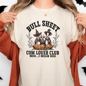 May include: A cream-colored t-shirt featuring a graphic of three cows wearing witch hats surrounded by pumpkins. The text reads BULL SHEET COW LOVER CLUB MOO...I MEAN BOO. The design is framed by thin black branches and foliage elements.