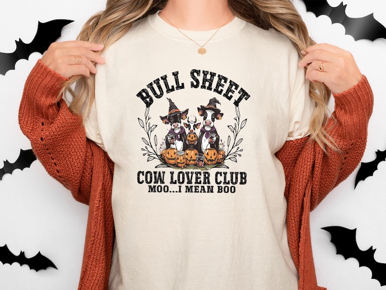 May include: A cream-colored t-shirt featuring a graphic of three cows wearing witch hats surrounded by pumpkins. The text reads BULL SHEET COW LOVER CLUB MOO...I MEAN BOO. The design is framed by thin black branches and foliage elements.