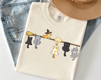 Może przedstawiać: Kremowy t-shirt z grafiką przedstawiającą siedem kotów z kreskówek wiszących na drewnianym trzonku miotły. Jeden pomarańczowy kot ma na głowie czarny kapelusz czarownicy. Koty są w różnych kolorach, w tym czarnym, szarym, pomarańczowym i białym w pomarańczowe łaty.