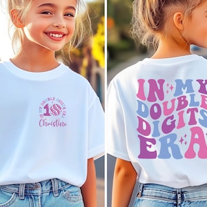 May include: White t-shirt with a colorful graphic design. The front features a pink and purple "10" with the text "In My Double Digits Era" and a name. The back of the shirt has the same text in a retro font.