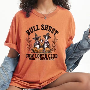 May include: An orange t-shirt featuring a graphic of three cows wearing witch hats surrounded by pumpkins. The text reads BULL SHEET COW LOVER CLUB MOO...I MEAN BOO. The shirt has short sleeves and a crew neckline, paired with a blue denim jacket.