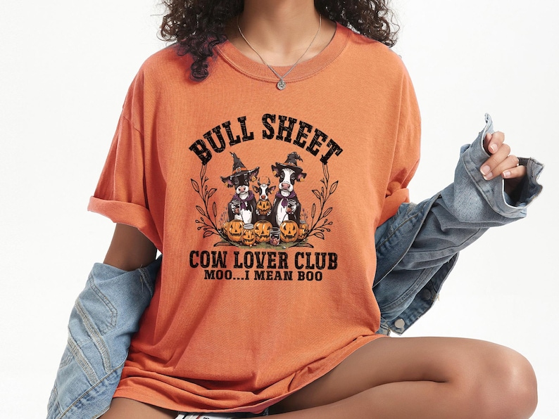 May include: An orange t-shirt featuring a graphic of three cows wearing witch hats surrounded by pumpkins. The text reads BULL SHEET COW LOVER CLUB MOO...I MEAN BOO. The shirt has short sleeves and a crew neckline, paired with a blue denim jacket.