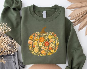 May include: An olive green sweatshirt featuring a floral pumpkin design. The pumpkin is composed of orange, yellow, and red flowers with a brown stem. The sweatshirt is folded, with the sleeve visible.