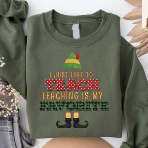 May include: Olive green sweatshirt with the text "I JUST LIKE TO TEACH TEACHING IS MY FAVORITE". The word "TEACH" is in red with white polka dots. The word "FAVORITE" is in green and black plaid. An elf hat and shoes are also featured.