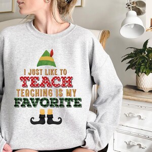 May include: A light gray sweatshirt with the text "I JUST LIKE TO TEACH TEACHING IS MY FAVORITE" in a festive design. The design includes an elf hat, red polka dot letters, and elf shoes.