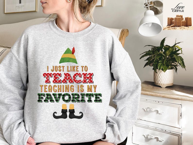 May include: A light gray sweatshirt with the text "I JUST LIKE TO TEACH TEACHING IS MY FAVORITE" in a festive design. The design includes an elf hat, red polka dot letters, and elf shoes.